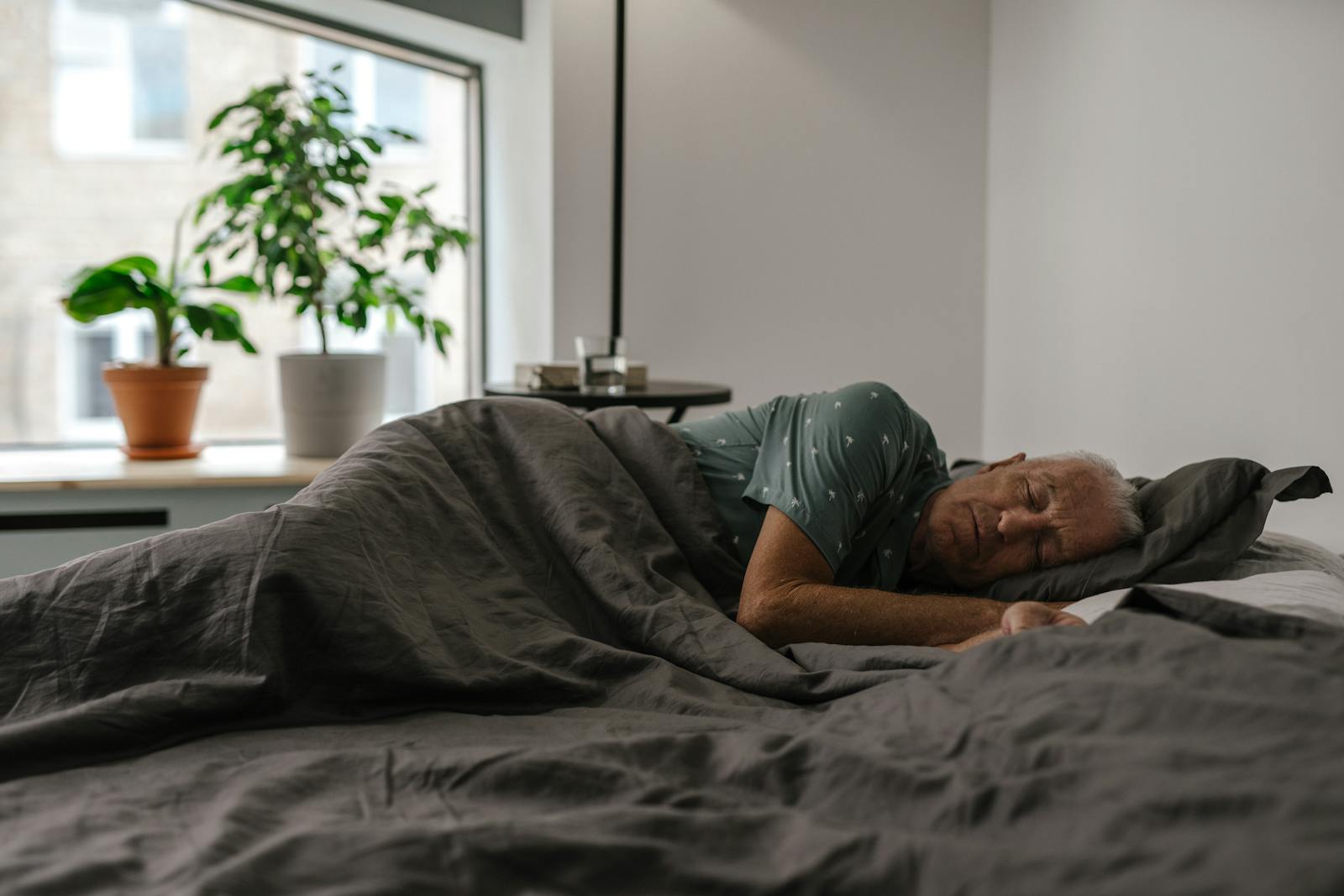 Senior man resting peacefully on a bed inside a room with natural light and plants.